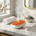 A white and orange heated pet grooming brush sits on a marble bathroom counter with steam rising from it, alongside a rolled white towel and a small succulent bowl.