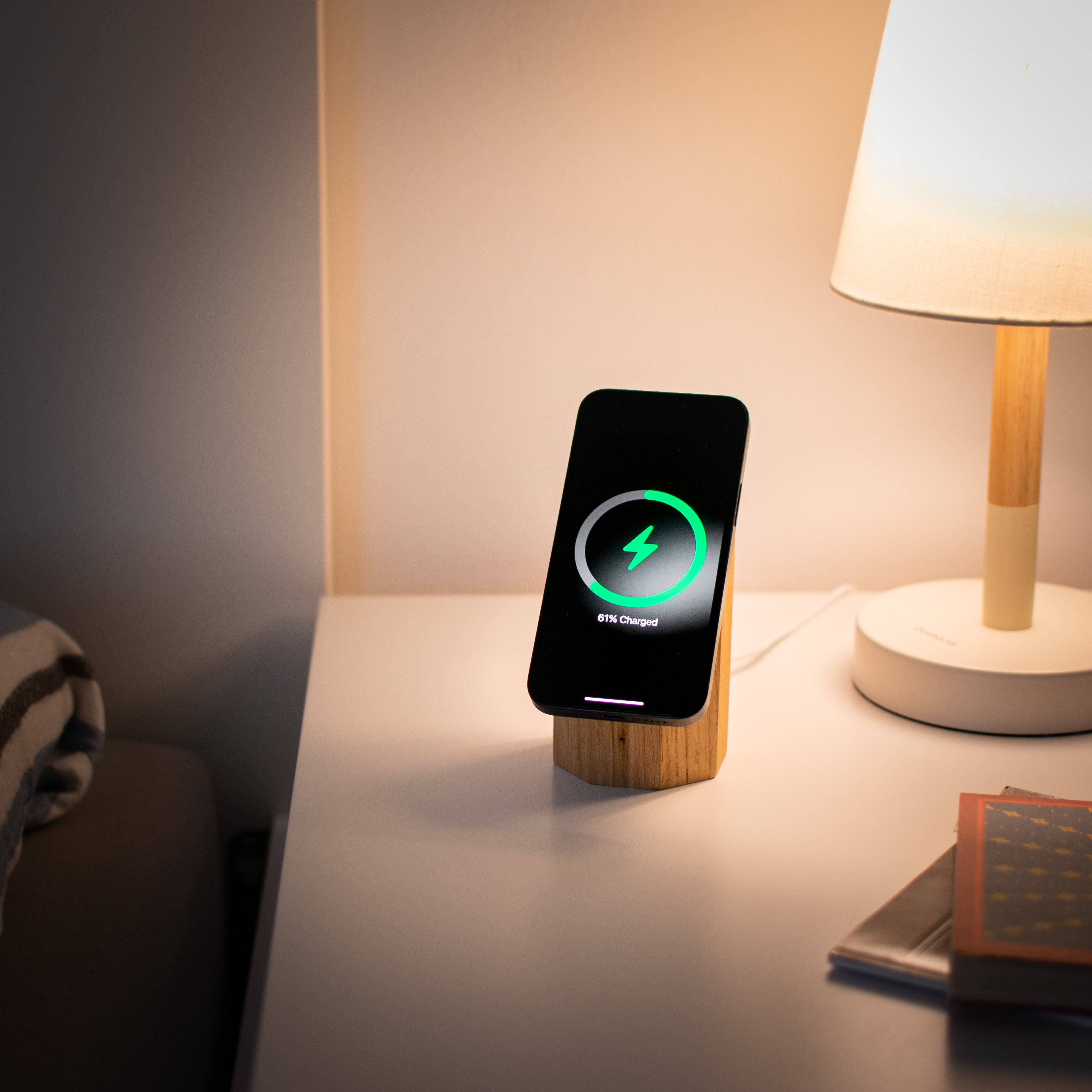 Smartphone on a wooden stand with charging indicator on a nightstand next to a lamp.